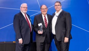 three men in business suits stand in a line shoulder to should and the middle man is holding a glass award